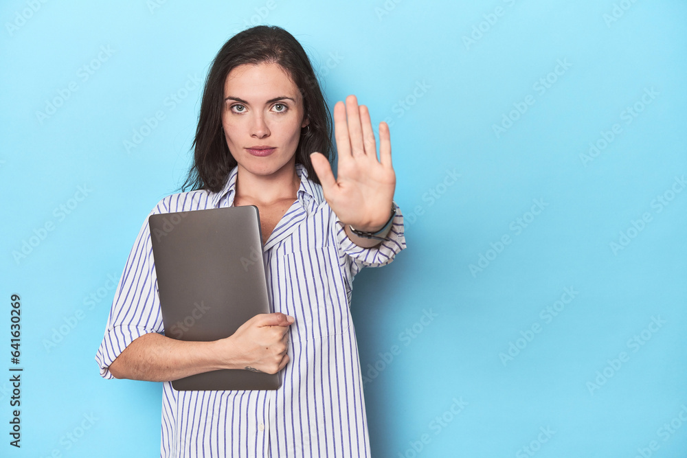 Young businesswoman holding laptop on blue standing with outstretched hand showing stop sign, preventing you. Young businesswoman holding laptop on blue standing with outstretched hand showing stop sign, preventing you.