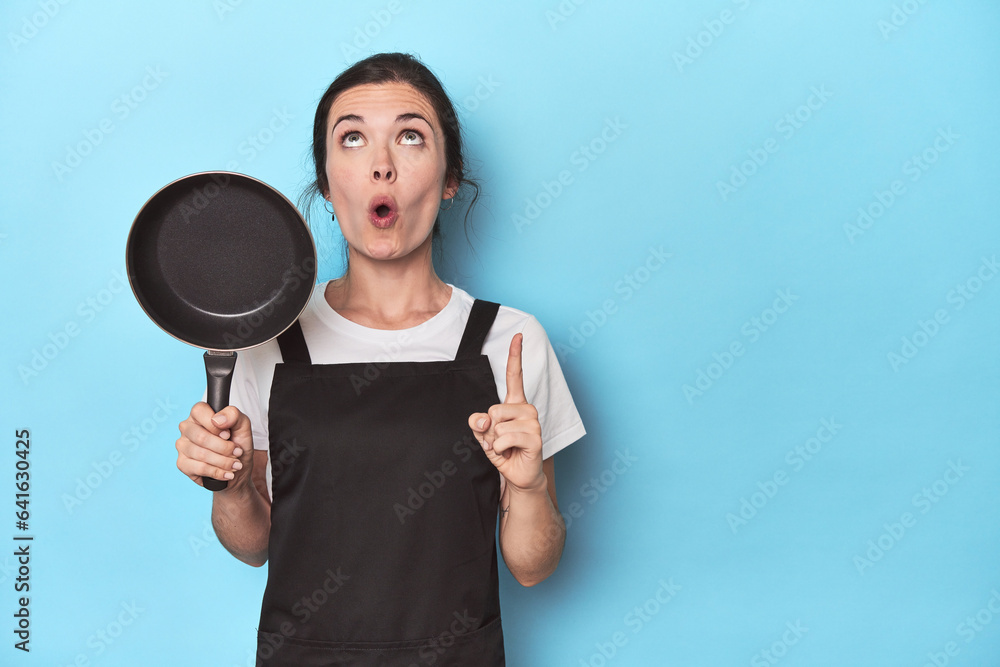 Woman with apron and pan on blue background pointing upside with opened mouth. Woman with apron and pan on blue background pointing upside with opened mouth.