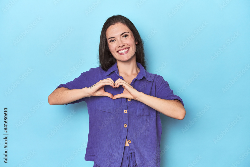Young caucasian woman on blue backdrop smiling and showing a heart shape with hands. Young caucasian woman on blue backdrop smiling and showing a heart shape with hands.
