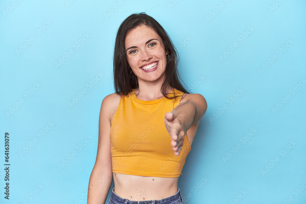 Fototapeta premium Young caucasian woman on blue backdrop stretching hand at camera in greeting gesture.