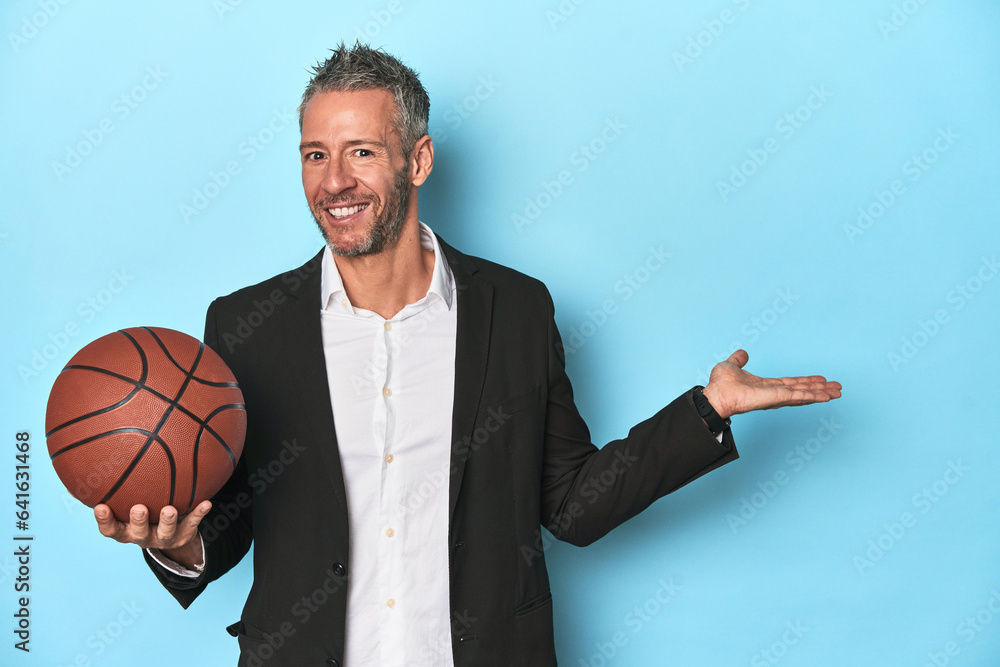 Middle-aged basketball coach on blue backdrop Middle-aged basketball coach on blue backdropshowing a copy space on a palm and holding another hand on waist.