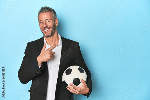 Caucasian football coach, middle-aged on blue set smiling and pointing aside, showing something at blank space.