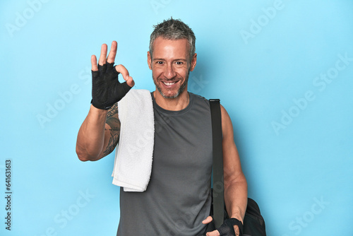 Athlete with gym backpack on blue background cheerful and confident showing ok gesture.
