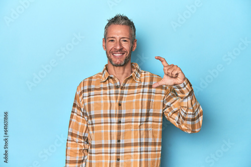 Middle-aged caucasian man on blue backdrop holding something little with forefingers, smiling and confident.