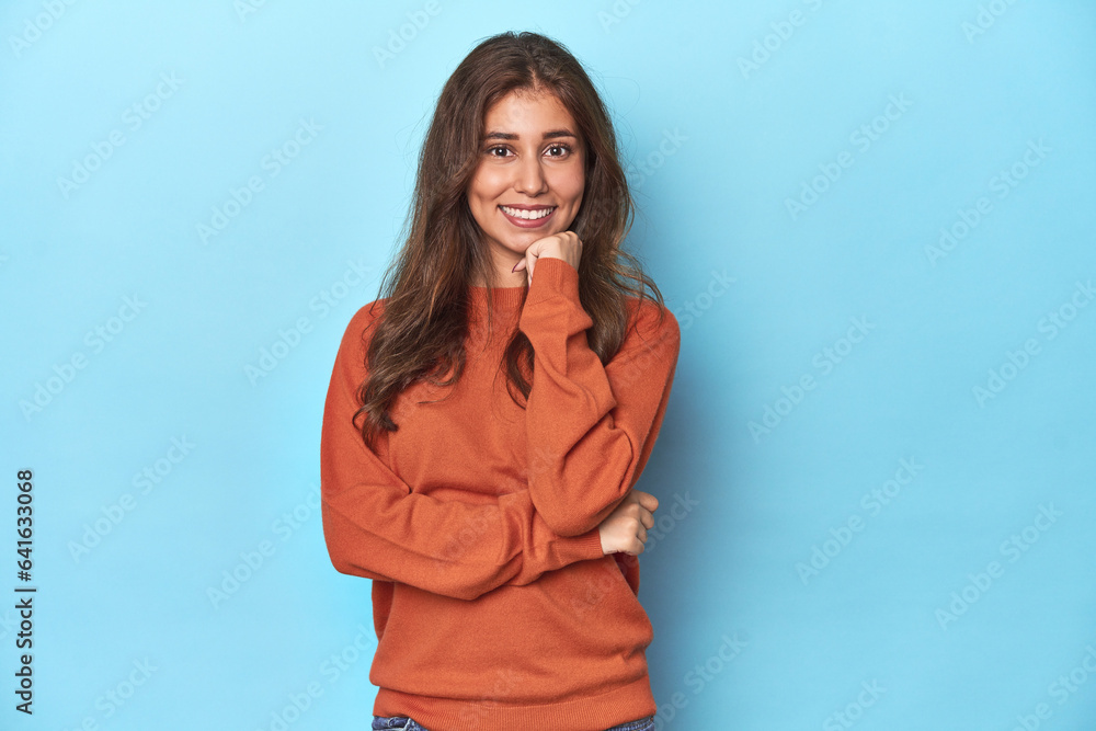 Fototapeta premium Teen girl in vibrant orange sweater on blue smiling happy and confident, touching chin with hand.