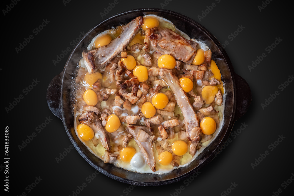 Large cast-iron frying pan with fried meat on the bones and yellow eggs on a black background. Large cast-iron frying pan with fried meat on the bones and yellow eggs on a black background.