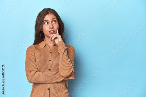 Teen girl in brown polo shirt on blue looking sideways with doubtful and skeptical expression.