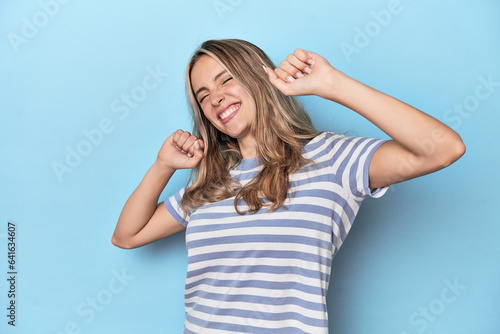 Blonde young caucasian woman in blue studio celebrating a special day, jumps and raise arms with energy.