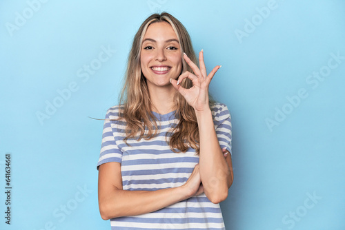 Blonde young caucasian woman in blue studio winks an eye and holds an okay gesture with hand.