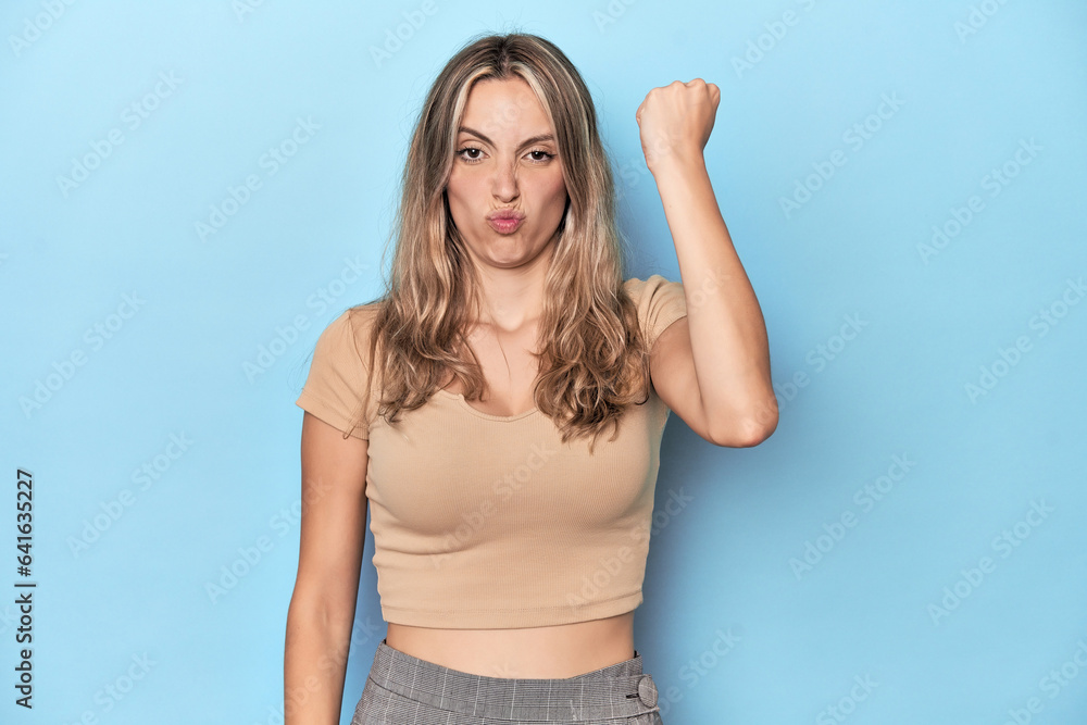 Blonde young caucasian woman in blue studio showing fist to camera, aggressive facial expression. Blonde young caucasian woman in blue studio showing fist to camera, aggressive facial expression.