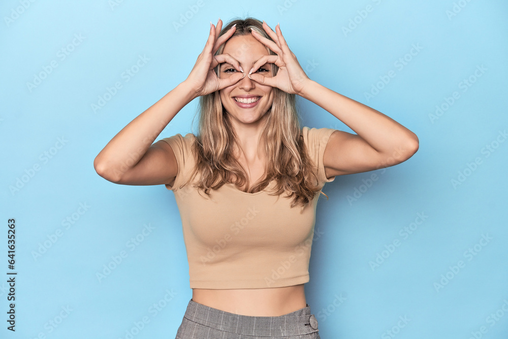 Blonde young caucasian woman in blue studio showing okay sign over eyes Blonde young caucasian woman in blue studio showing okay sign over eyes