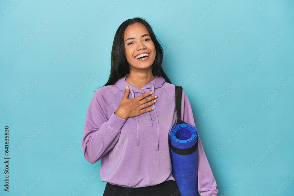 Filipina athlete with yoga mat on blue laughs out loudly keeping hand on chest. Filipina athlete with yoga mat on blue laughs out loudly keeping hand on chest.