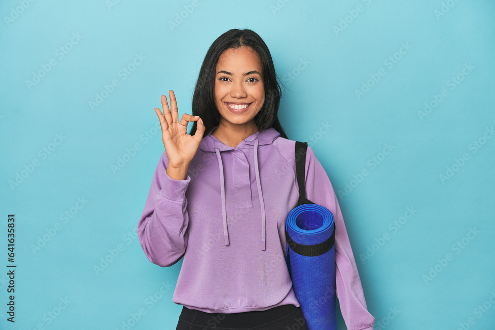Filipina athlete with yoga mat on blue cheerful and confident showing ok gesture. Filipina athlete with yoga mat on blue cheerful and confident showing ok gesture.