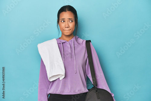 Filipina with gym gear on blue studio confused, feels doubtful and unsure.