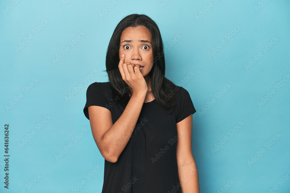 Filipina young woman on blue studio biting fingernails, nervous and very anxious.