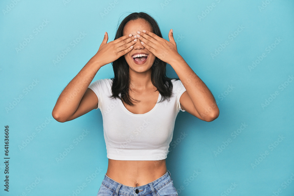 Filipina young woman on blue studio covers eyes with hands, smiles broadly waiting for a surprise. Filipina young woman on blue studio covers eyes with hands, smiles broadly waiting for a surprise.