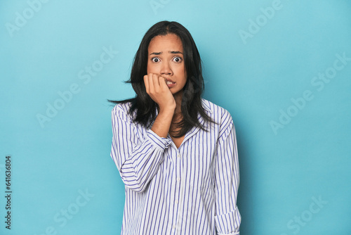 Filipina young woman on blue studio biting fingernails, nervous and very anxious.