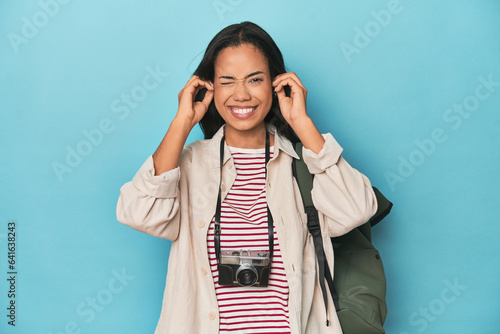 Filipina woman with camera and backpack on blue covering ears with hands.