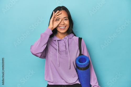 Filipina athlete with yoga mat on blue excited keeping ok gesture on eye.