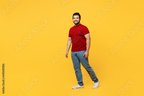 Full body side profile view young cheerful fun happy Indian man he wearing red t-shirt casual clothes walking going looking camera isolated on plain yellow orange background studio. Lifestyle concept.