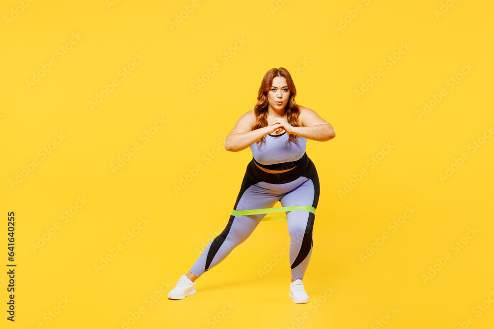 Full body young chubby plus size big fat fit woman wear blue top warm up train use rubber elastic bands for legs do squats isolated on plain yellow background studio home gym. Workout sport concept.