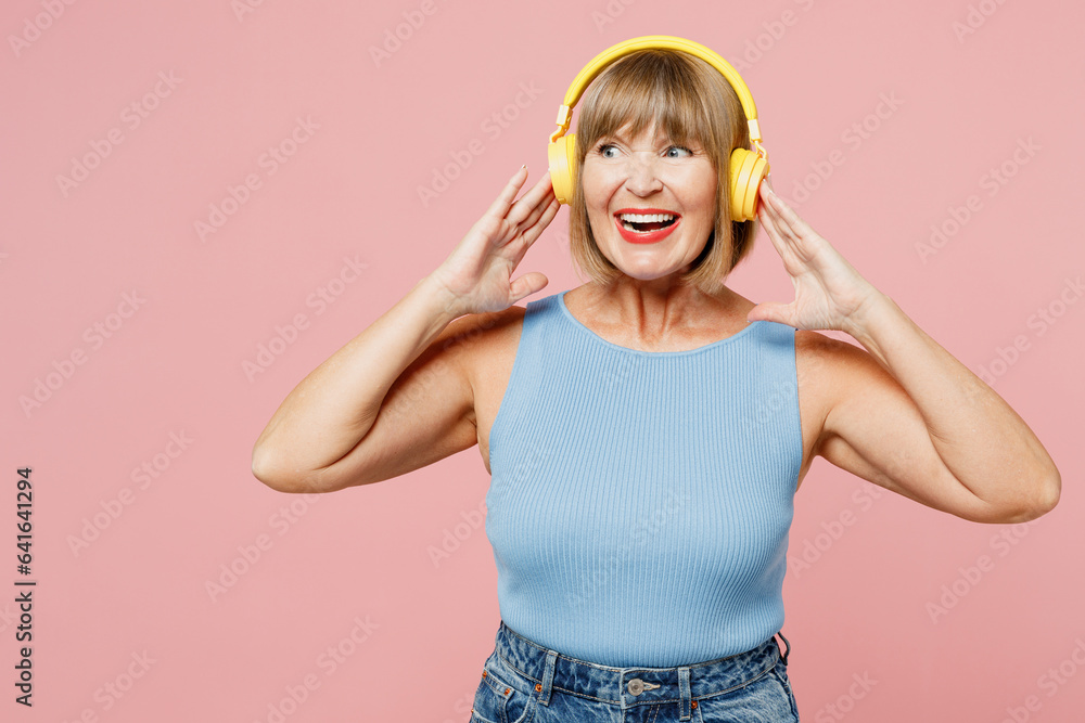 Elderly blonde woman 50s years old she wears blue undershirt casual clothes listen to music on headphones look aside on area isolated on plain pastel light pink background studio. Lifestyle concept. Elderly blonde woman 50s years old she wears blue undershirt casual clothes listen to music on headphones look aside on area isolated on plain pastel light pink background studio. Lifestyle concept.