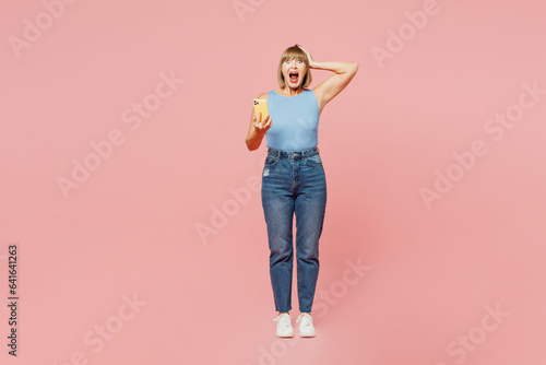 Full body surprised excited elderly blonde woman 50s years old wears blue undershirt casual clothes hold head use mobile cell phone isolated on plain pastel light pink background. Lifestyle concept.