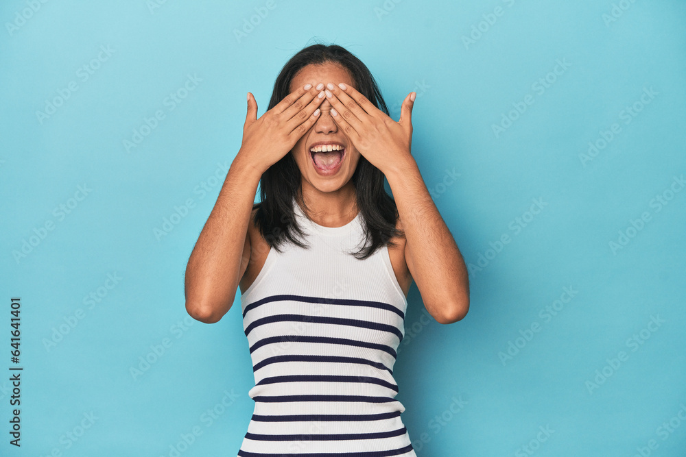 Filipina young woman on blue studio covers eyes with hands, smiles broadly waiting for a surprise. Filipina young woman on blue studio covers eyes with hands, smiles broadly waiting for a surprise.