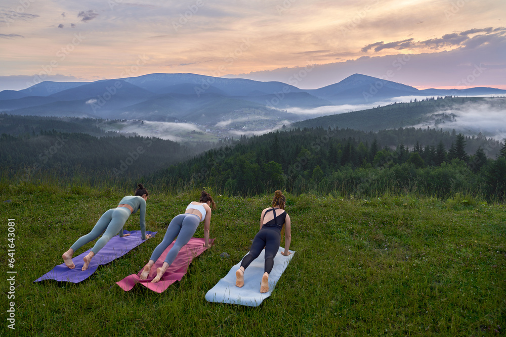 Naklejka premium Active young women in sport clothes doing yoga exercises on fresh air. Three fit ladies enjoying outdoors training during summer sunset.