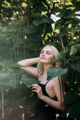 A positive pretty woman of age with long blond hair, in a black long sexy dress, stands and relaxes among the bushes with large green leaves in the city park.