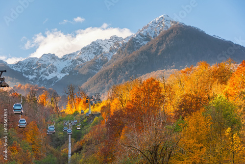 Autumn landscape with mountain range green  yellow red foliage of forest trees illuminated by the sun. High mountains with snow capped peaks with cable car  and passenger cabins rising up.