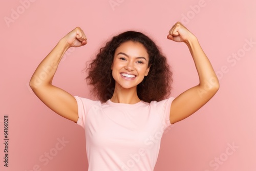 Medium shot portrait photography of a grinning girl in her 20s making a i'm strong gesture showing muscles against a pastel pink background. With generative AI technology © Markus Schröder
