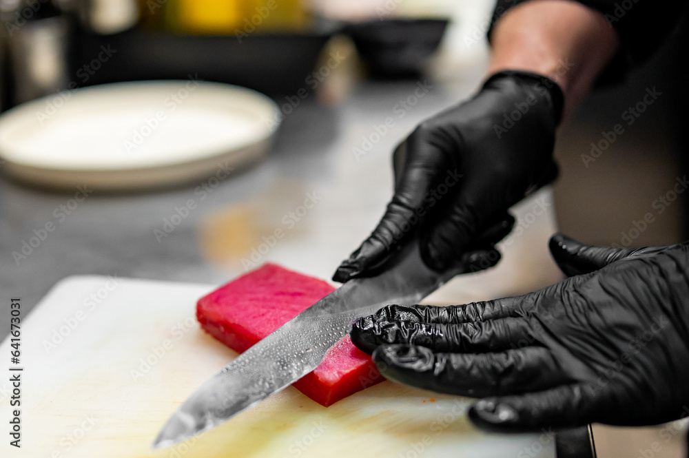Fototapeta premium Close-up of hand sushi chef in gloves slices fresh tuna fish fillet at sushi bar.