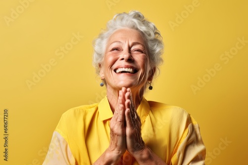 Headshot portrait photography of a joyful mature woman joining palms in a gesture of gratitude against a pastel yellow background. With generative AI technology