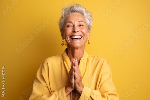 Headshot portrait photography of a joyful mature woman joining palms in a gesture of gratitude against a pastel yellow background. With generative AI technology