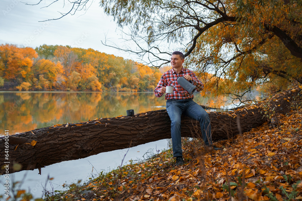 a man sits on a log on the river bank and drinks tea or coffee from a thermos, enjoys the landscape, beautiful nature in the autumn season