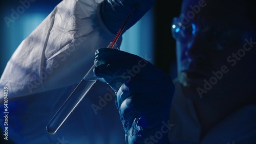 A forensic investigator adds a blood sample to a test tube with a reagent using a pipette. A man collects evidence at the crime scene, in a dark apartment lit by blue police sirens. photo