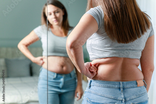 Konstfotografi Displeased young woman touching her belly looking in mirror at home