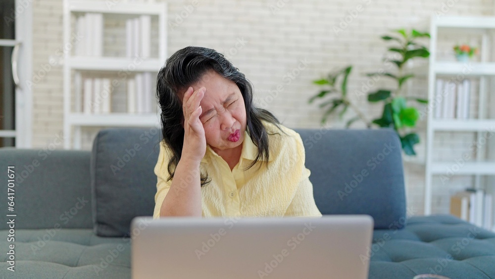 Asian elderly woman having headache migraine while working on laptop sitting in sofa at living room. Tired stressed upset senior woman having headache. Health problem concept Asian elderly woman having headache migraine while working on laptop sitting in sofa at living room. Tired stressed upset senior woman having headache. Health problem concept