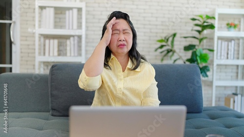 Asian elderly woman having headache migraine while working on laptop sitting in sofa at living room. Tired stressed upset senior woman having headache. Health problem concept