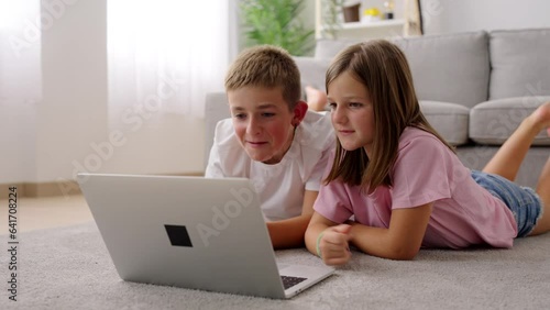 Two preteen children watching a movie on laptop lying on the floor. Brother and sister having fun