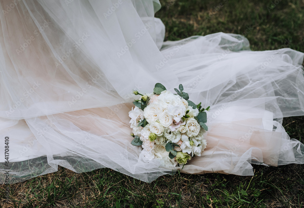 Fototapeta premium A beautiful bouquet of flowers, roses lies on a white dress with a long veil. Close-up wedding photography.
