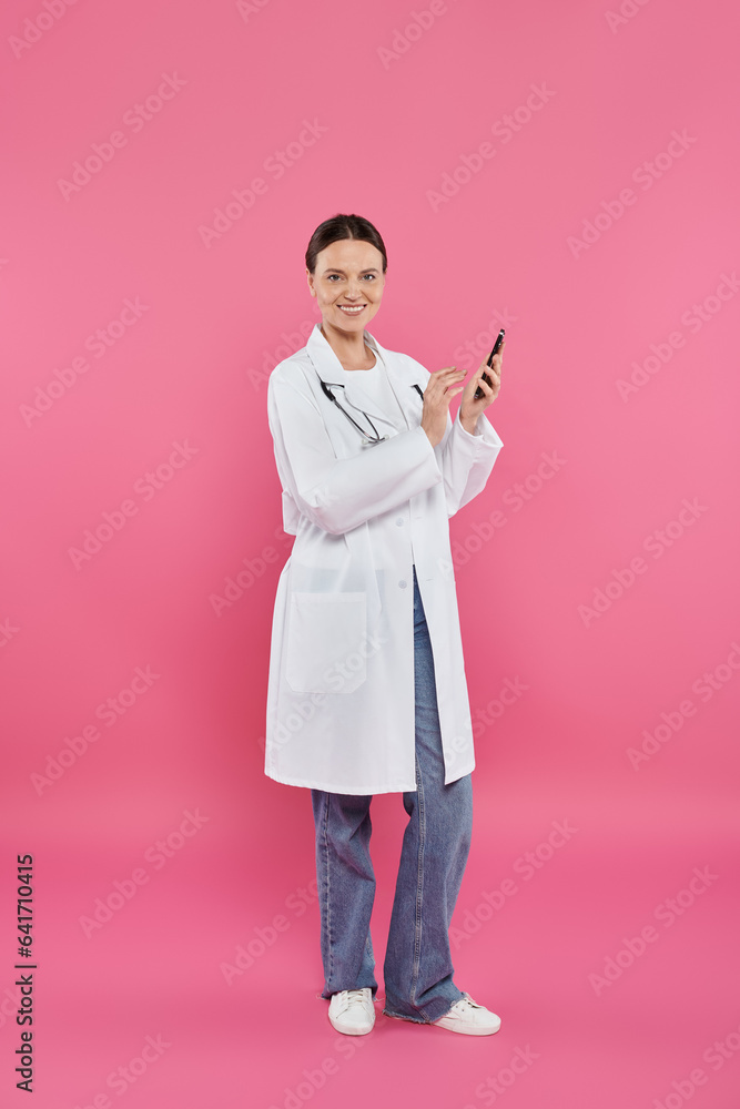 smiling doctor in white coat holding smartphone and looking at camera on pink, breast cancer concept smiling doctor in white coat holding smartphone and looking at camera on pink, breast cancer concept
