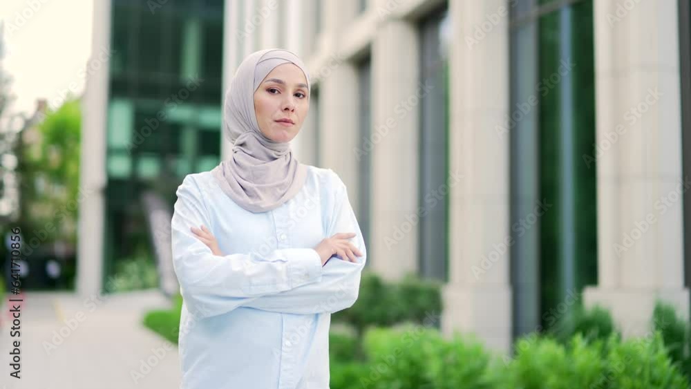 Portrait of a young serious muslim woman in hijab with crossed arms standing on the street near an office building. Head shot of a confident female manager or employee posing looking at the camera Portrait of a young serious muslim woman in hijab with crossed arms standing on the street near an office building. Head shot of a confident female manager or employee posing looking at the camera