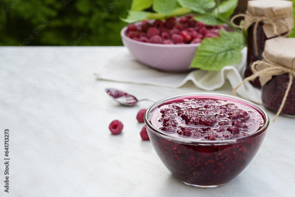 Bowl of raspberry jam and fresh berries with leaves on a white marble table with blurred green background. Banner, menu, recipe. Side view, place for text copy space. Bowl of raspberry jam and fresh berries with leaves on a white marble table with blurred green background. Banner, menu, recipe. Side view, place for text copy space.