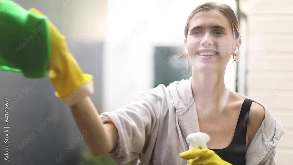 Young smiling woman washing window with green sponge. Happy beautiful girl cleaning window by spraying cleaning products and wiping with sponge in yellow gloves, earphones, send blow kiss and dance. Young smiling woman washing window with green sponge. Happy beautiful girl cleaning window by spraying cleaning products and wiping with sponge in yellow gloves, earphones, send blow kiss and dance.