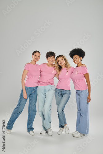 breast cancer awareness, cheerful multicultural women with pink ribbons hugging on grey, diversity