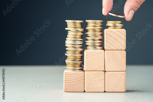Hand add a few coins at the top of the final level built by wood cubes as step stairs, paying less at the long term, reduce cost, save for future, insurance, and investment, or subsidy money concept photo