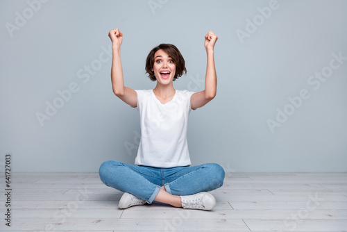 Full length photo of overjoyed girl wear stylish t-shirt rising hands up win gambling sit on floor isolated on gray color background