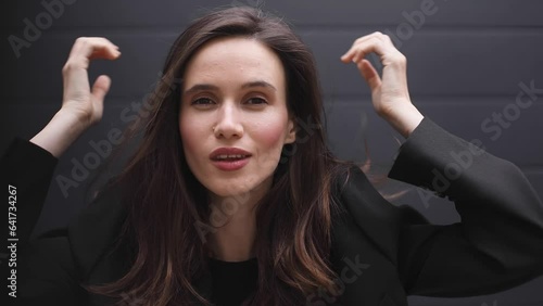 Lovely coquettish brunette woman wear black business jacket, winking playfully, flirting. Woman fix her hair isolated on black background. Curly hair fashion model face strong, smiling. Look at camera